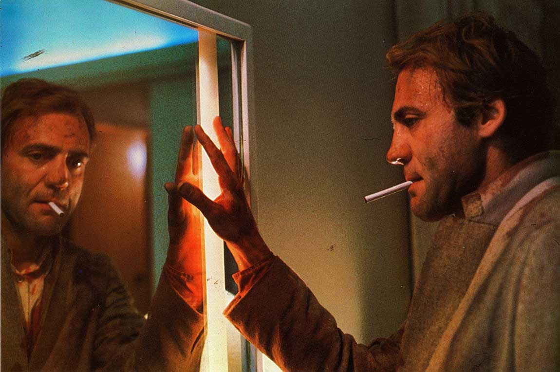 un homme avec une cigarette au bec,qui touche de sa main droite son reflet dans un miroir